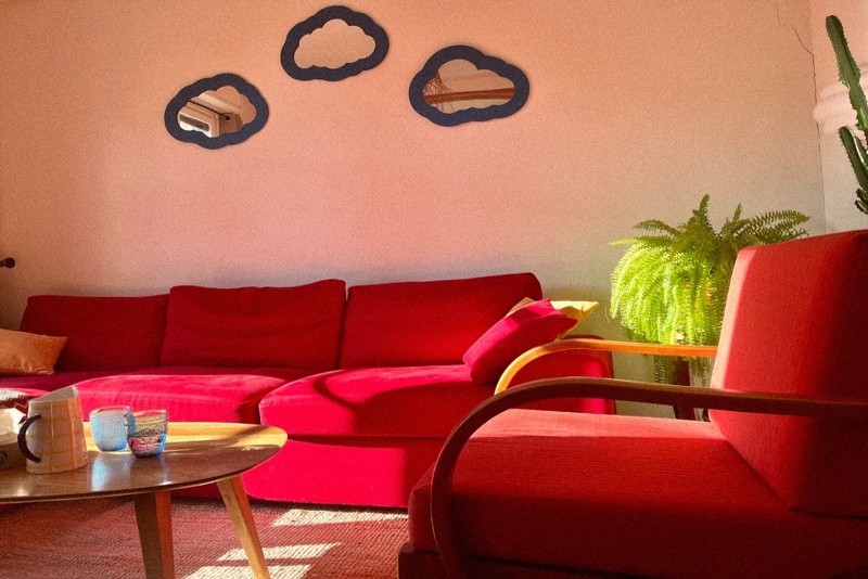 Salon d’un cabinet de consultation avec canapé rouge vif, fauteuil assorti, miroirs en forme de nuage et plantes vertes, baigné de lumière naturelle.